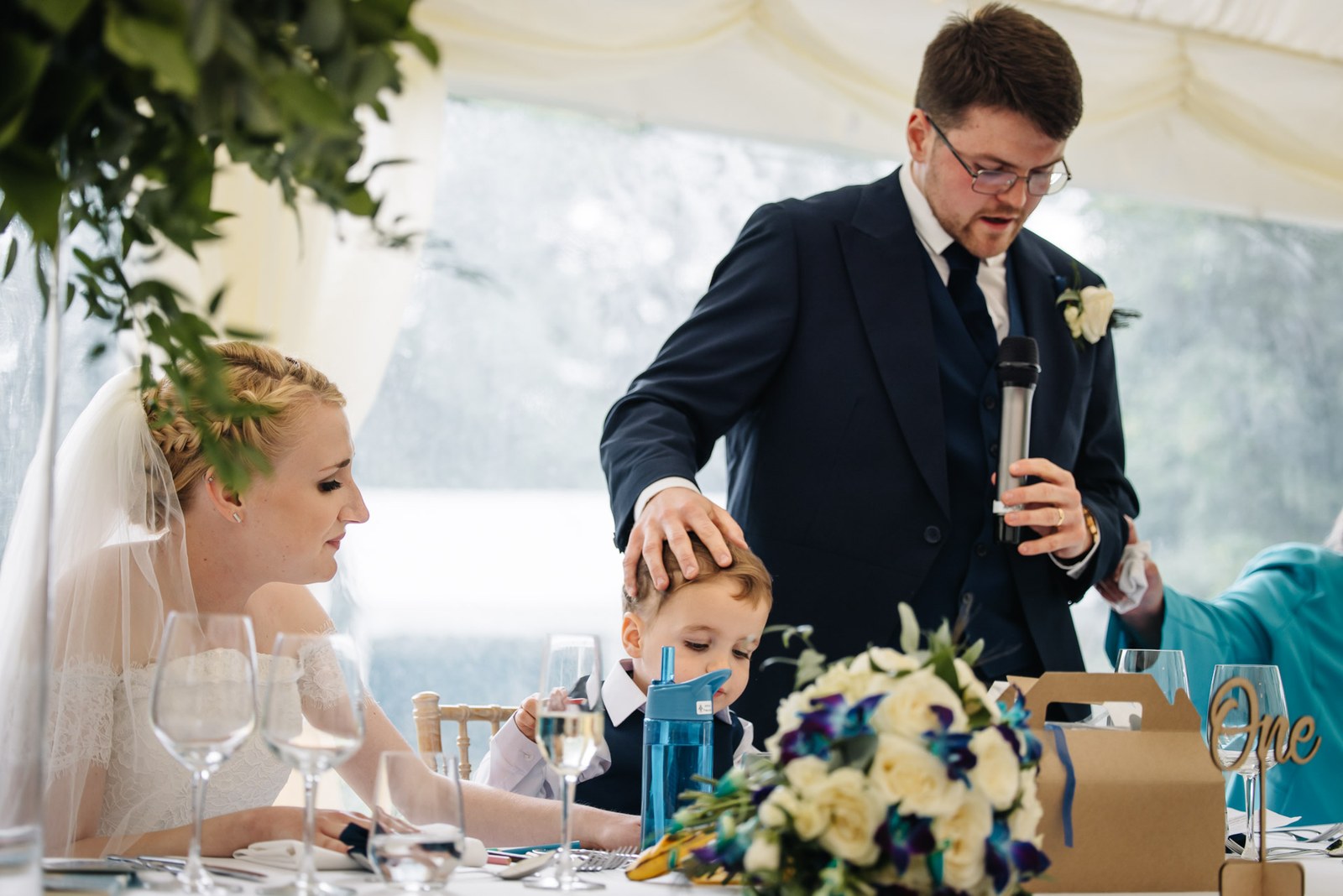 The groom giving his speech, a hand on the page boy's head, the bride seated