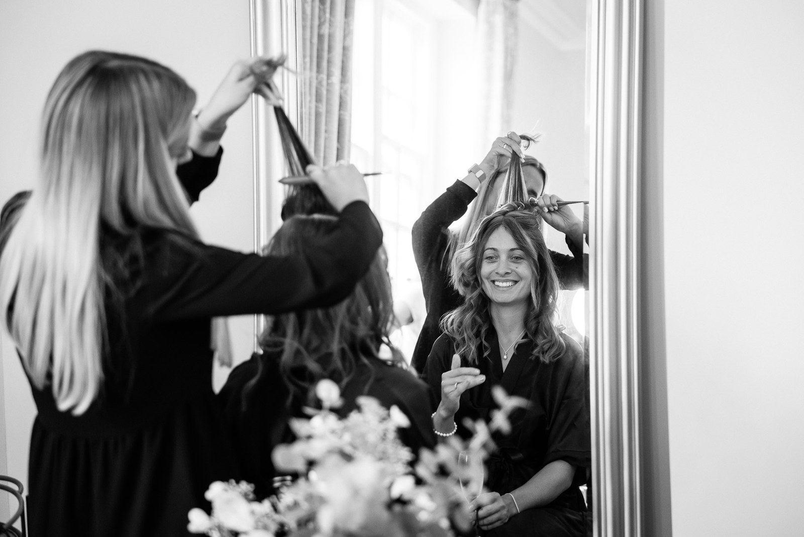 Black-and-white of the bride having her hair styled at a window during preparations