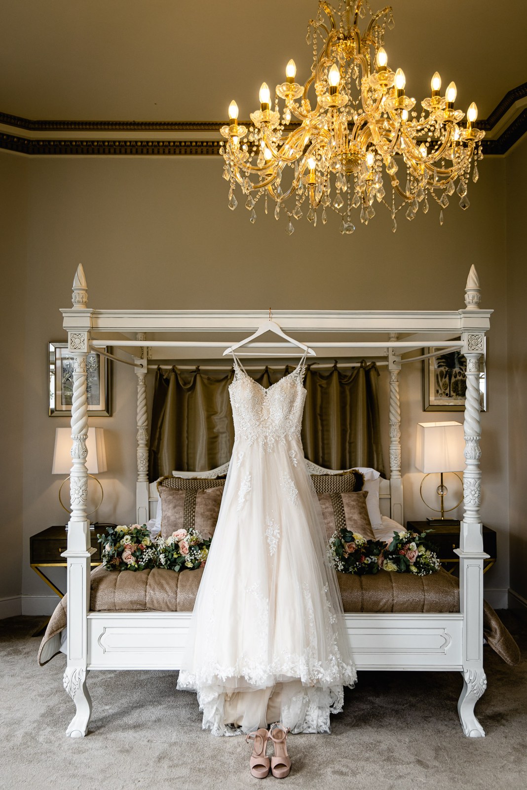 The bride's lace gown hanging on a four-poster bed beneath a chandelier at Bourton Hall