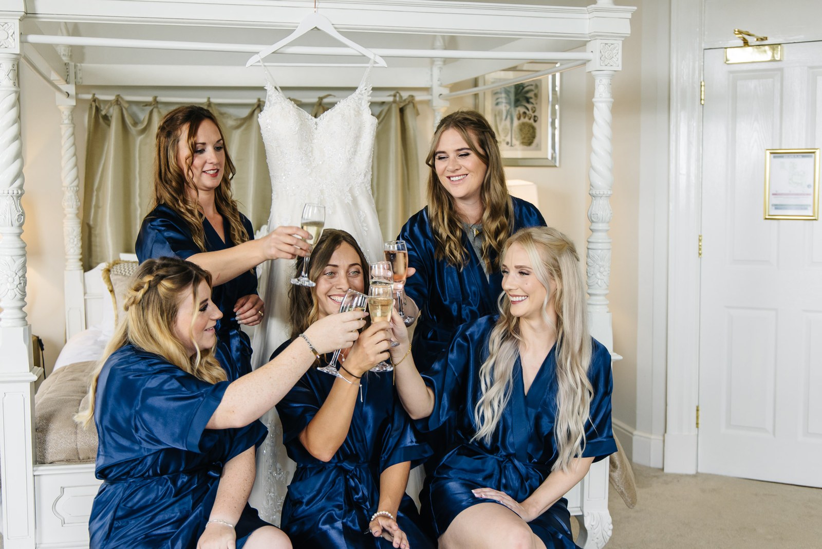 The bride and her bridesmaids in navy robes toasting with champagne beneath the hanging gown