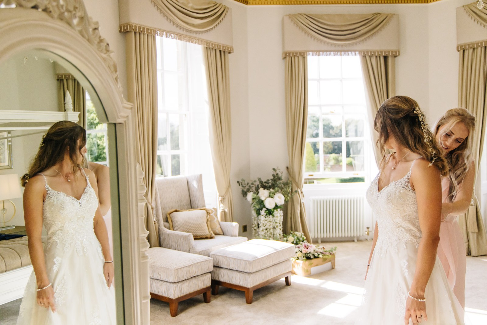 The bride in her gown beside a mirror as a bridesmaid helps in a sunlit room
