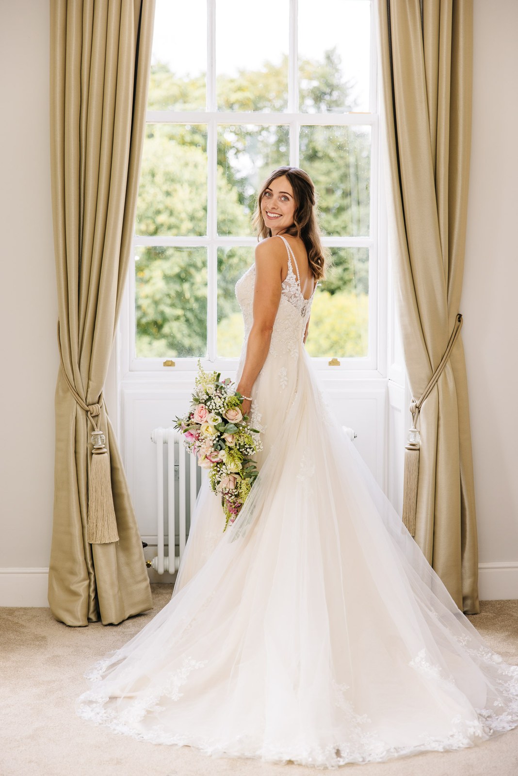 The bride posing with her bouquet at a tall curtained window at Bourton Hall