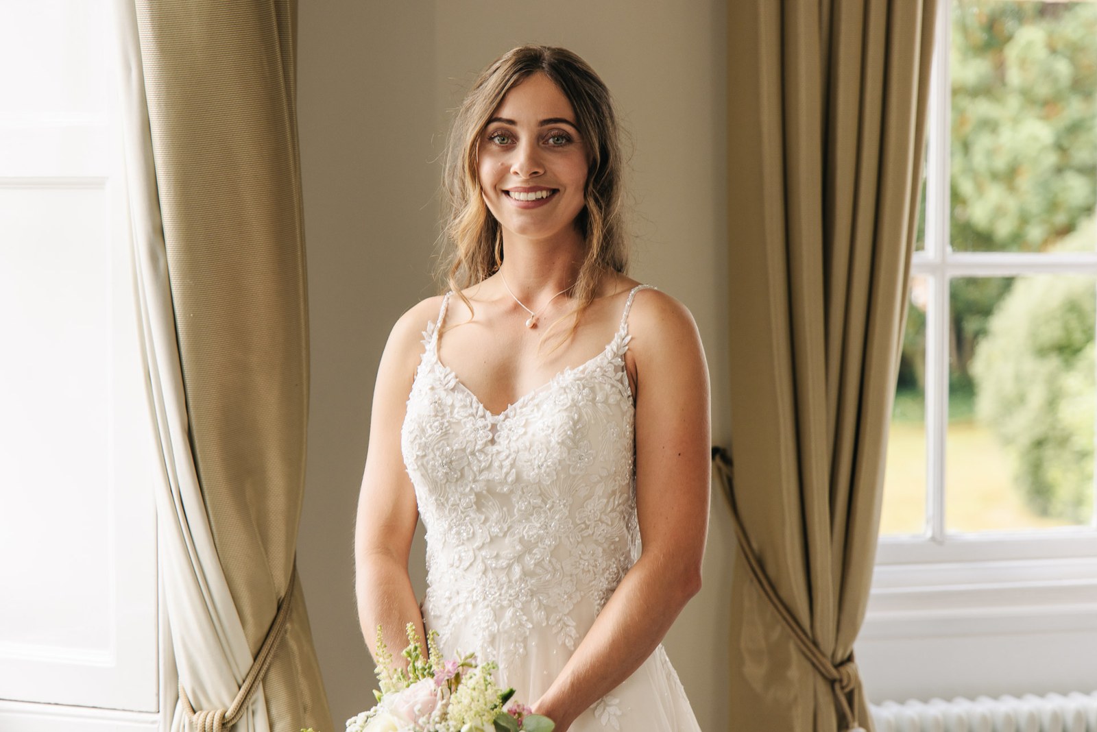 The bride smiling by the window in her lace gown, bouquet in hand