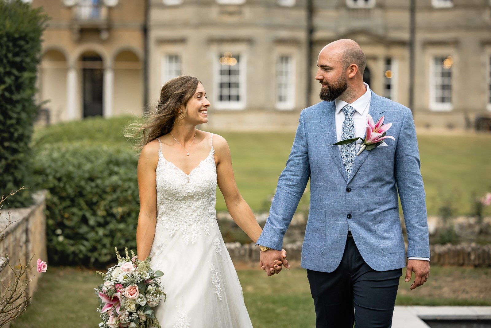 Bride and groom walking hand in hand on the lawn, Bourton Hall behind