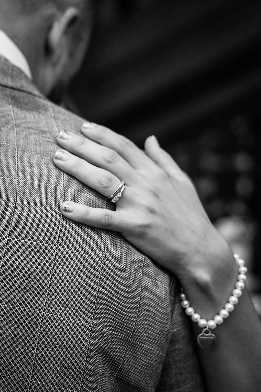 Black-and-white detail of the bride's ringed hand and pearl bracelet on the groom's shoulder