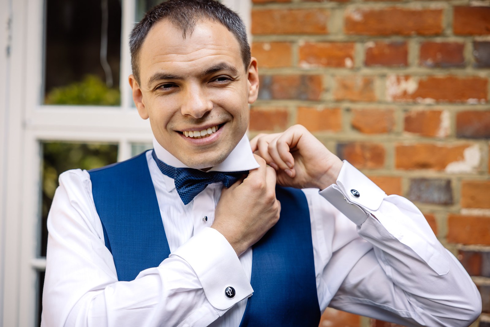 The groom adjusting his bow tie against a brick wall