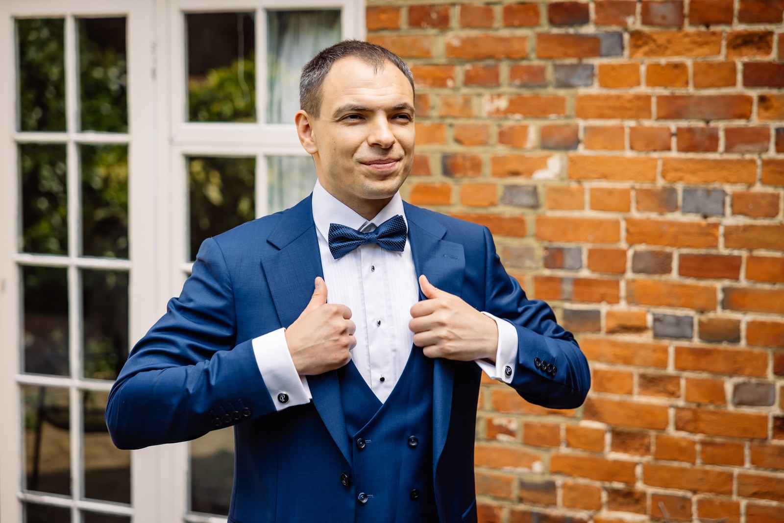 The groom straightening his blue jacket beside a brick wall