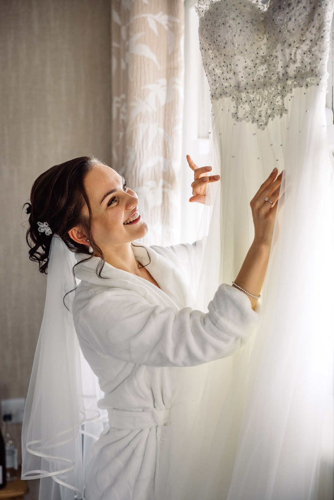 The bride in her robe reaching to touch her hanging dress at the window