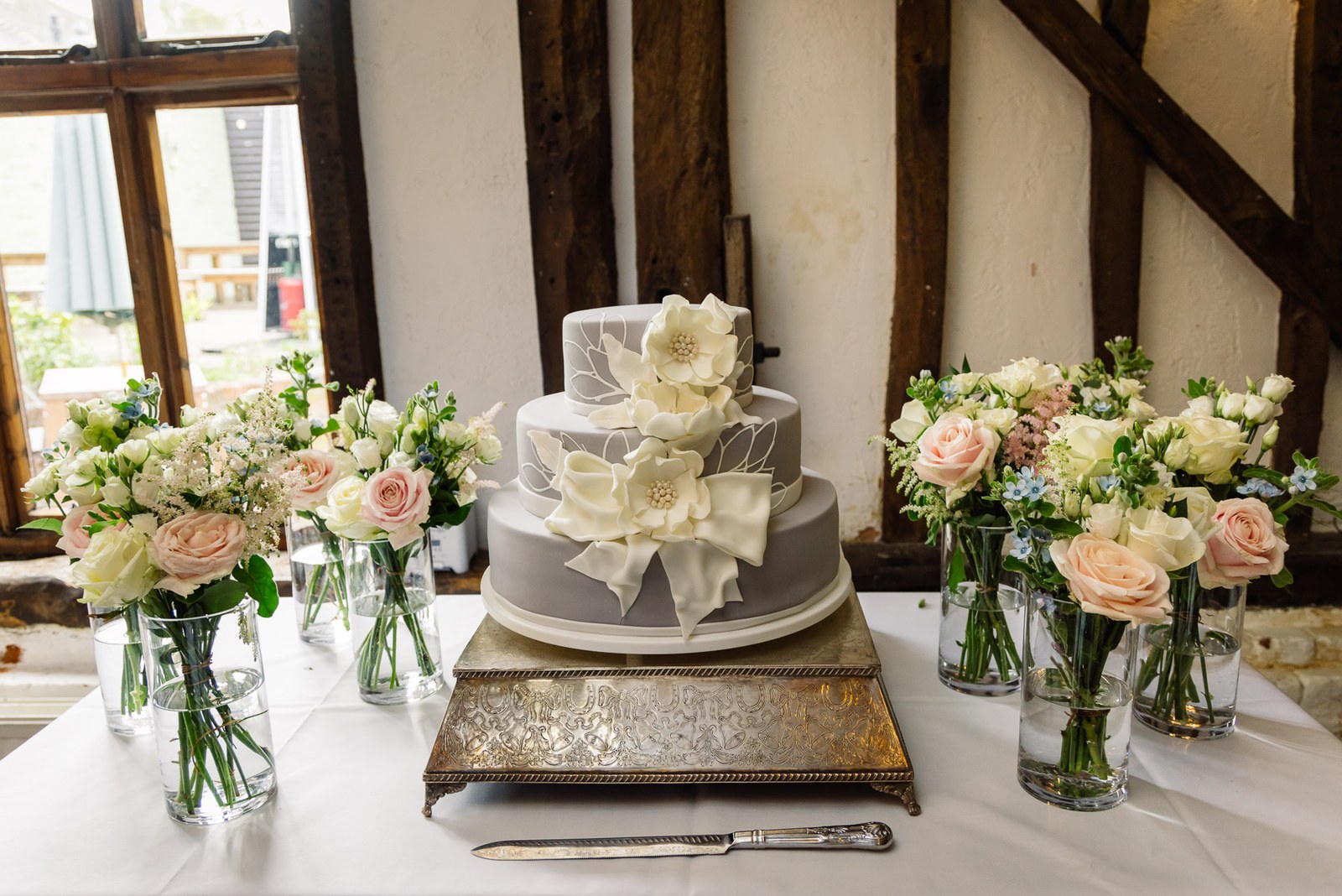 The tiered grey cake with a sugar flower among rose vases in the beamed room at Cantley House Hotel