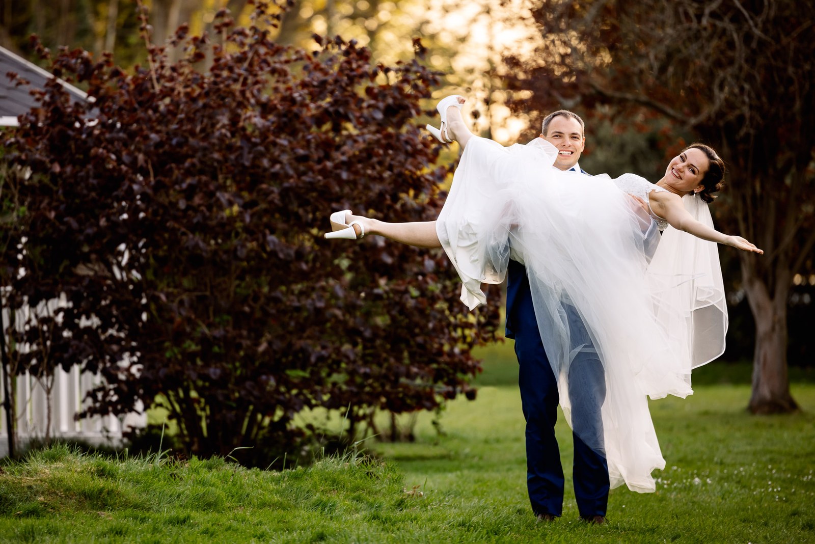 The groom holding the bride aloft, arms outstretched, before a copper beech