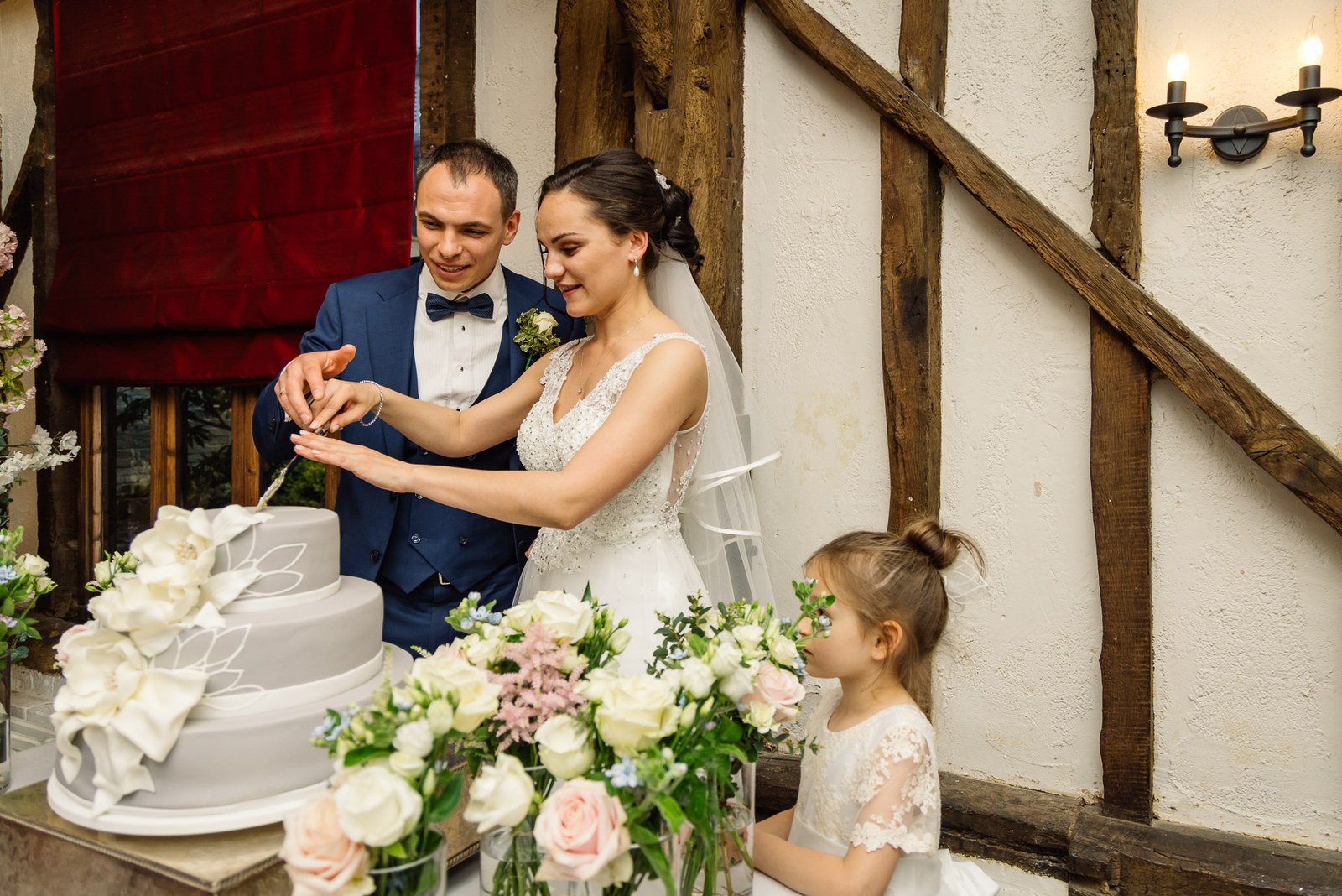 The couple cutting their cake in the beamed room, a flower girl beside them