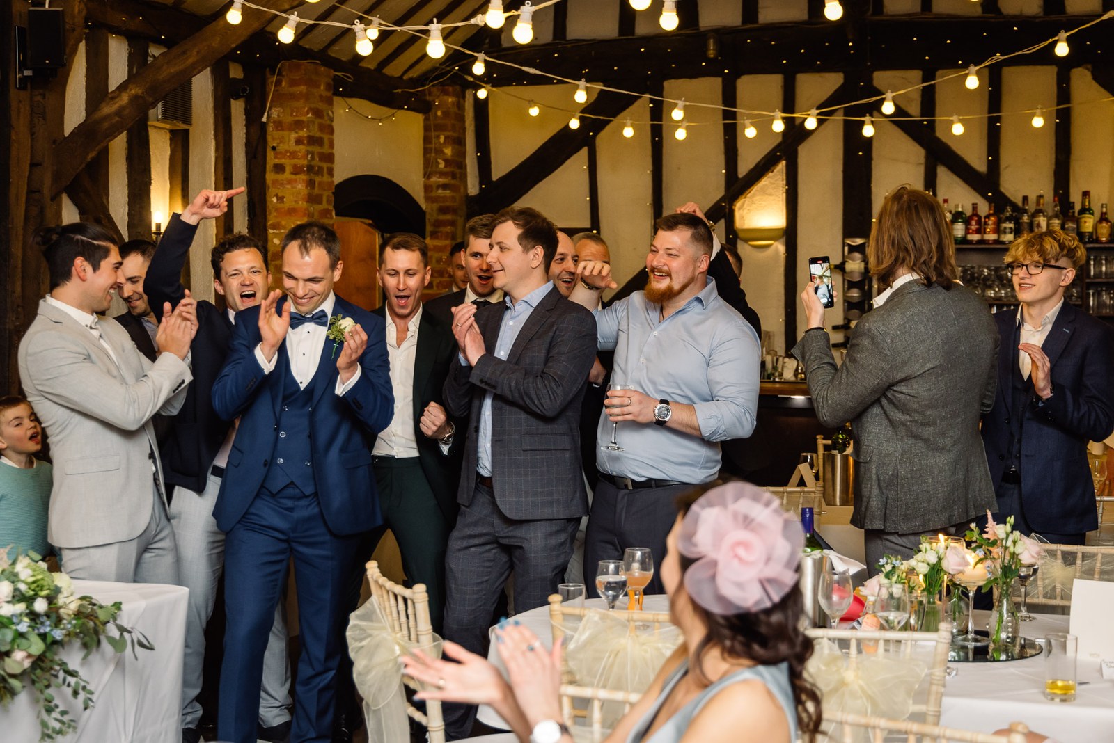 Groomsmen cheering under the festoon lights of the beamed reception at Cantley House Hotel