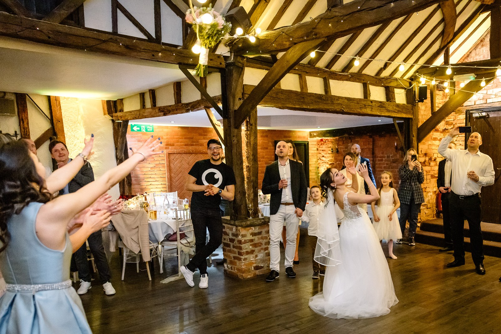 Guests reaching up for the bouquet the bride has just tossed in the beamed hall