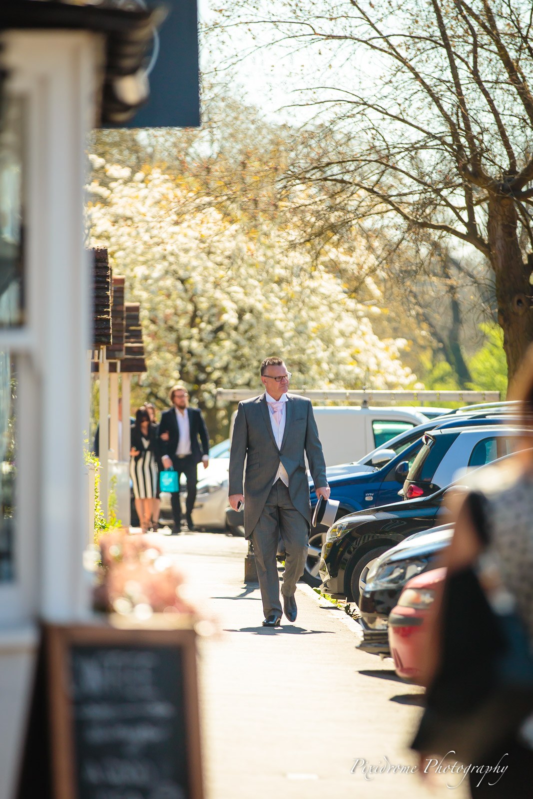 The groom in top hat and tails walking through the car park past spring blossom