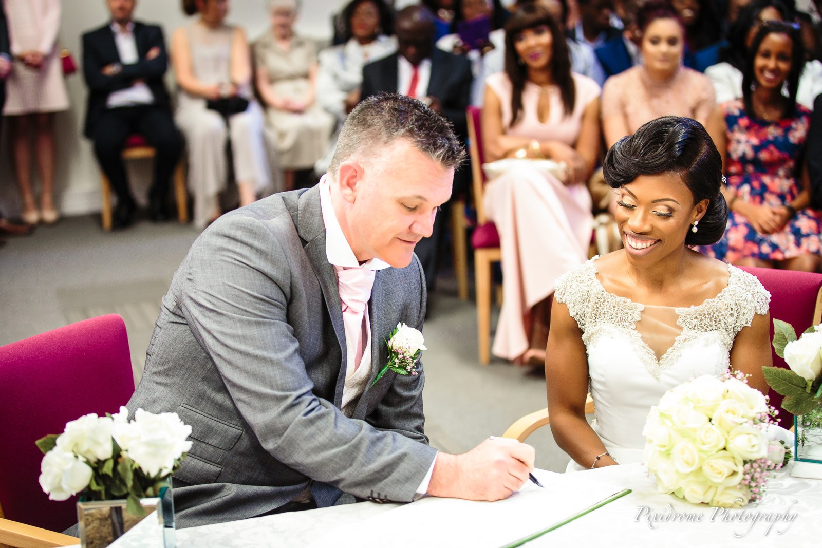 The groom signing the register as the bride looks on delightedly