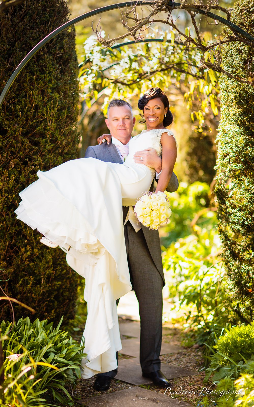 The groom carrying the bride through a blossom arch in the gardens of Chenies Manor House