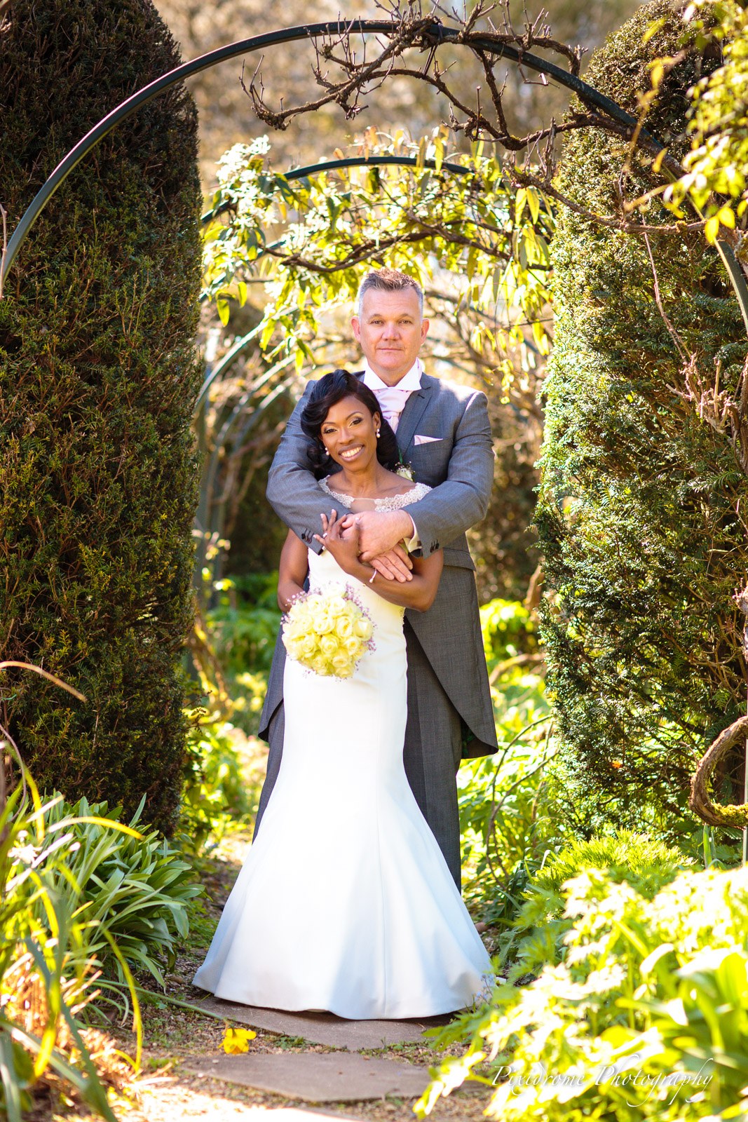 The couple embracing under a garden arch at Chenies Manor House, yellow bouquet in hand