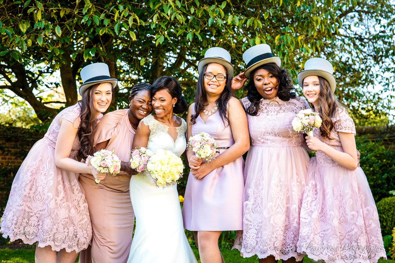 The bride with her bridesmaids in pink, some in grey top hats, beneath a tree