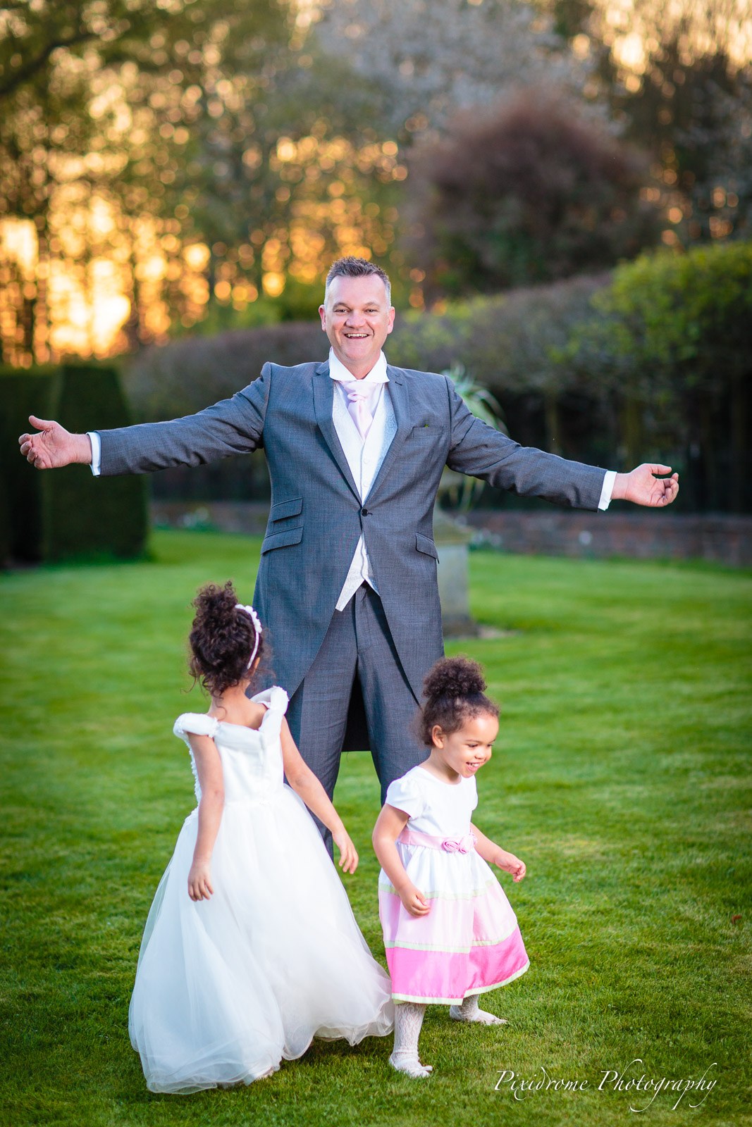 The groom opening his arms as two flower girls dance on the Chenies Manor House lawn