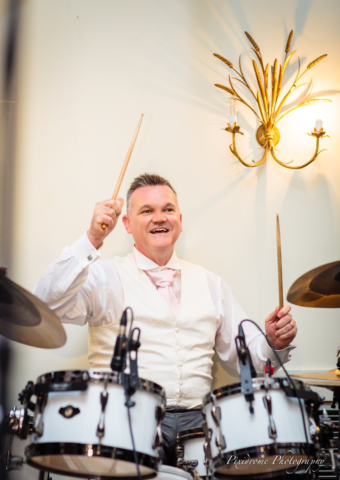 The groom playing the drums, sticks raised, mid-laugh