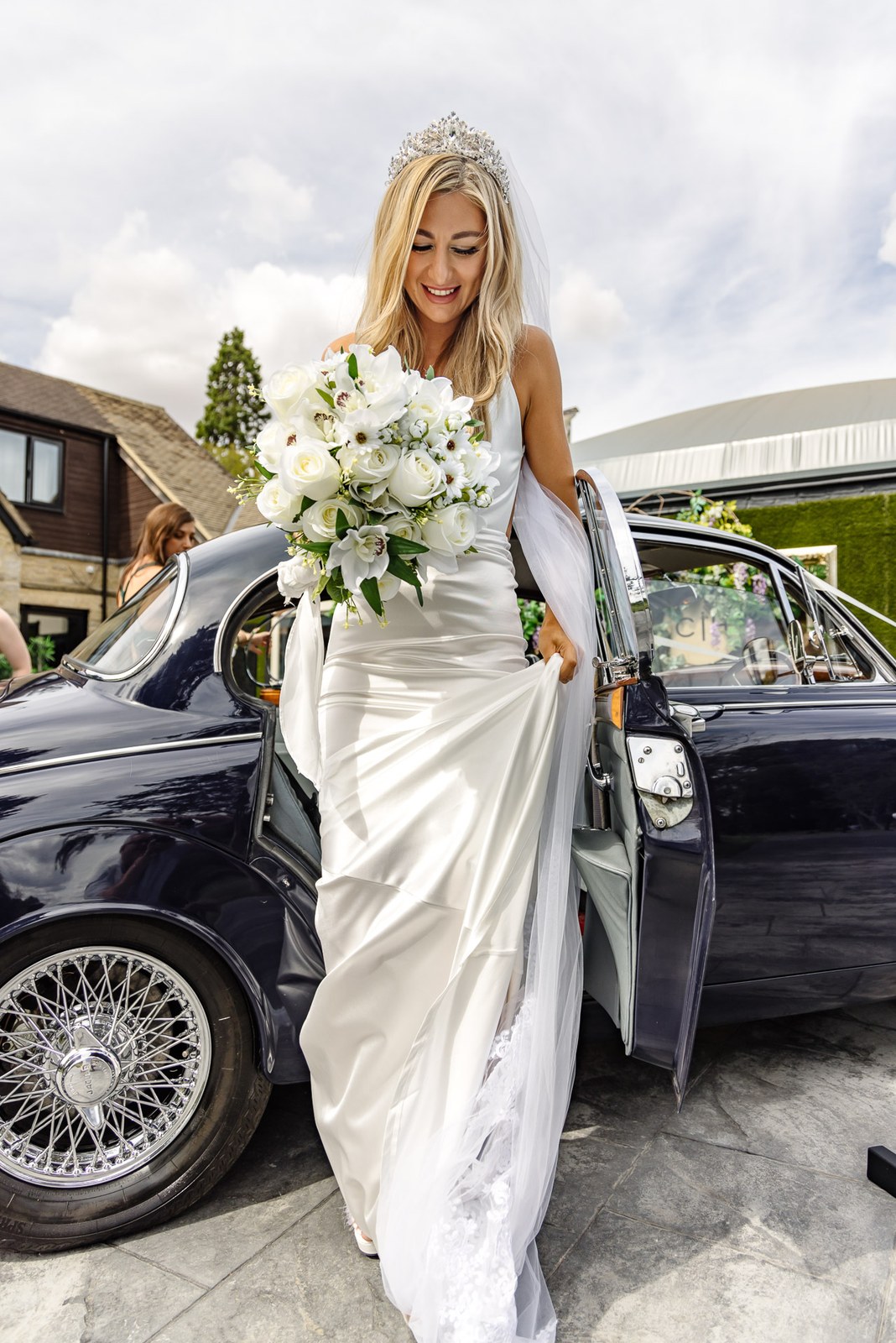 The bride stepping from the vintage car in her satin gown and tiara