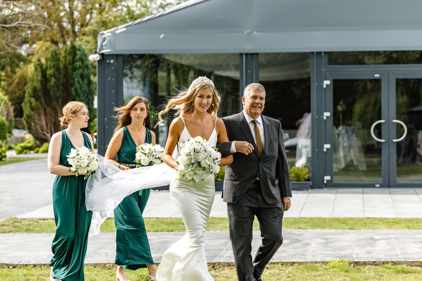 The bride arriving on her father's arm, the glass garden room of the Chesterton Hotel behind
