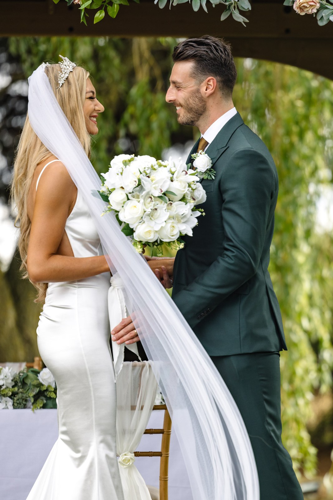 The couple smiling at each other under the flower arch, veil flowing