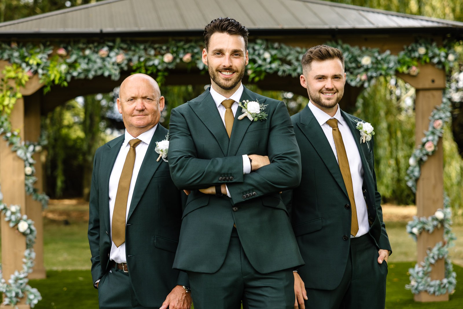 The groom with the best man and a groomsman beneath the foliage arch