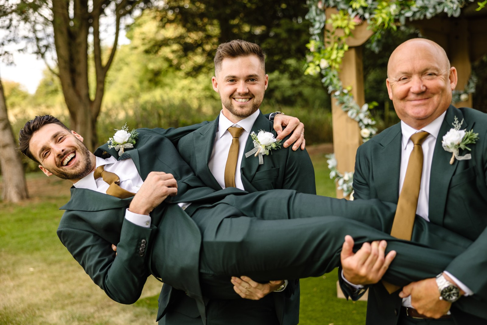 The best man hoisted horizontally by the groom and a groomsman on the lawn