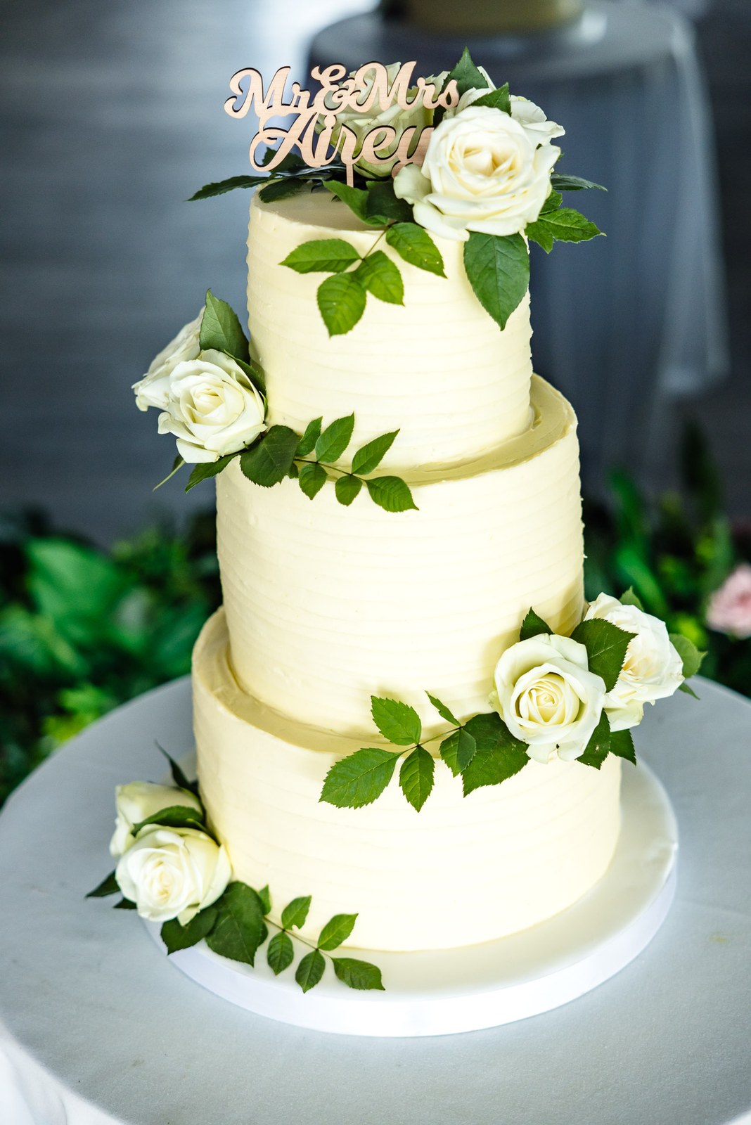 The three-tier cake dressed with white roses and foliage on a lit stand