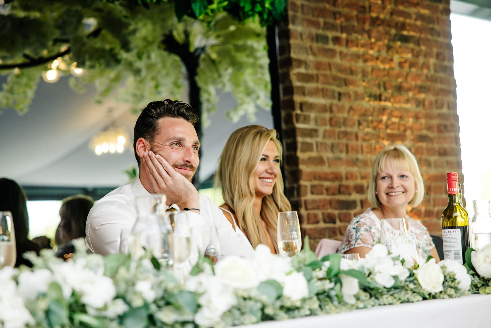 The groom resting his cheek on his hand, the bride and a guest beside him