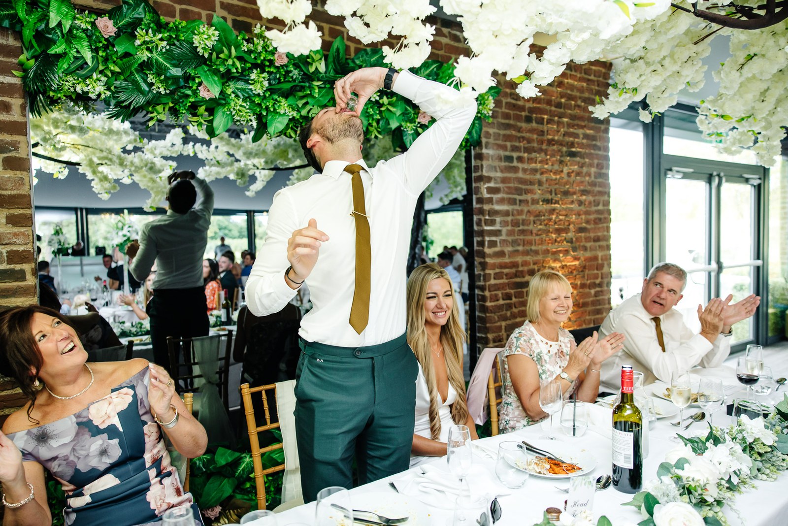 The groom downing a shot at the top table as guests clap and laugh