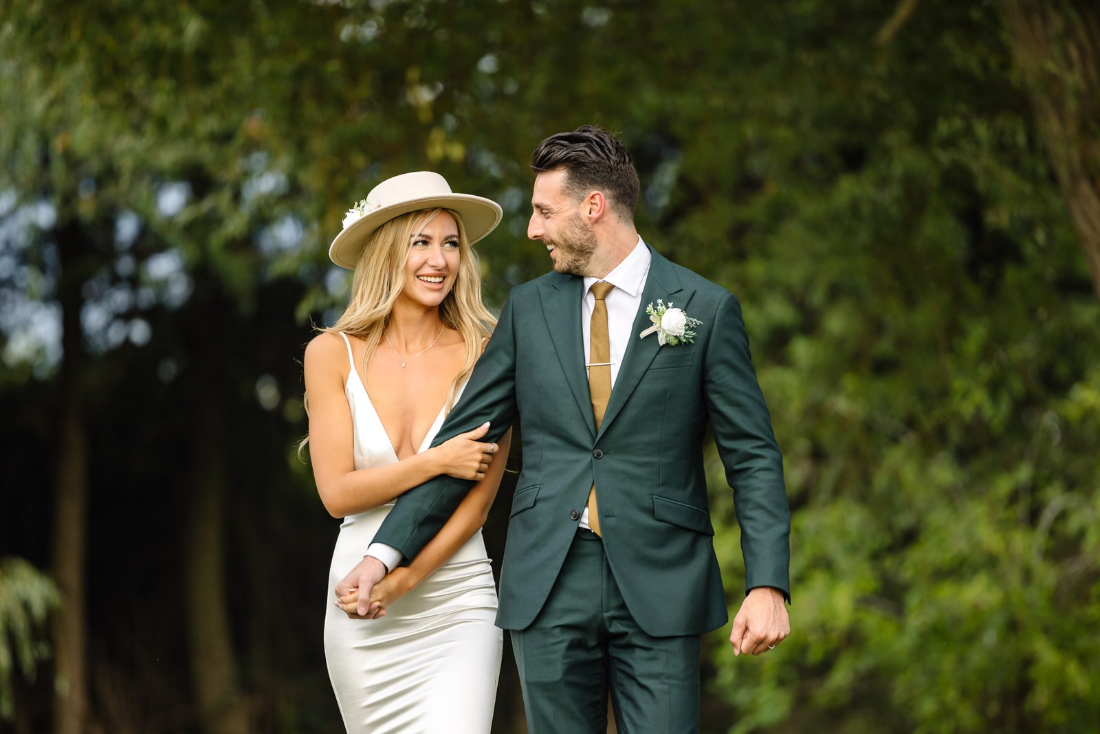 The couple walking among the trees at the Chesterton Hotel, the bride in a cream fedora