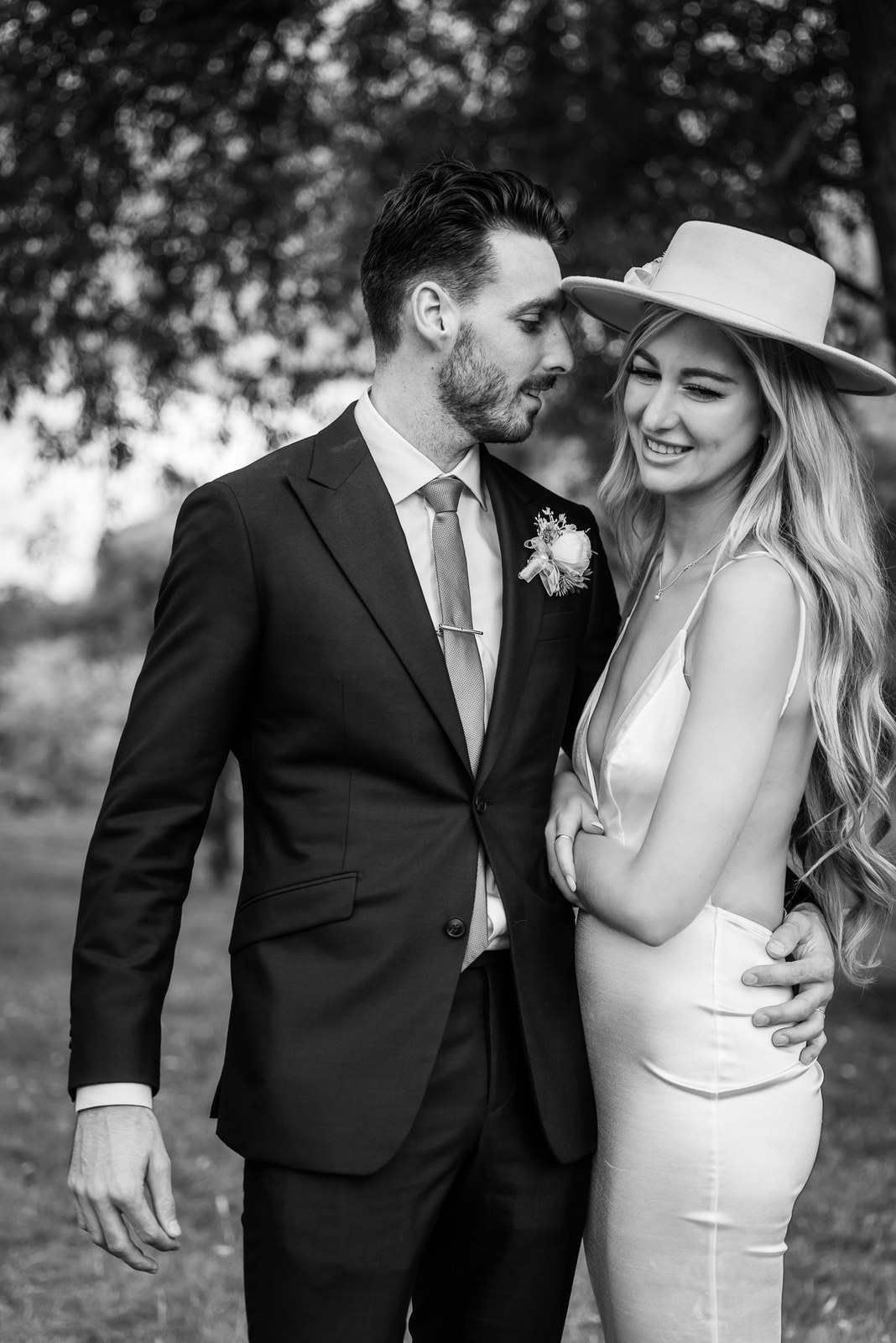 Black-and-white of the couple embracing under the trees, the bride in a fedora