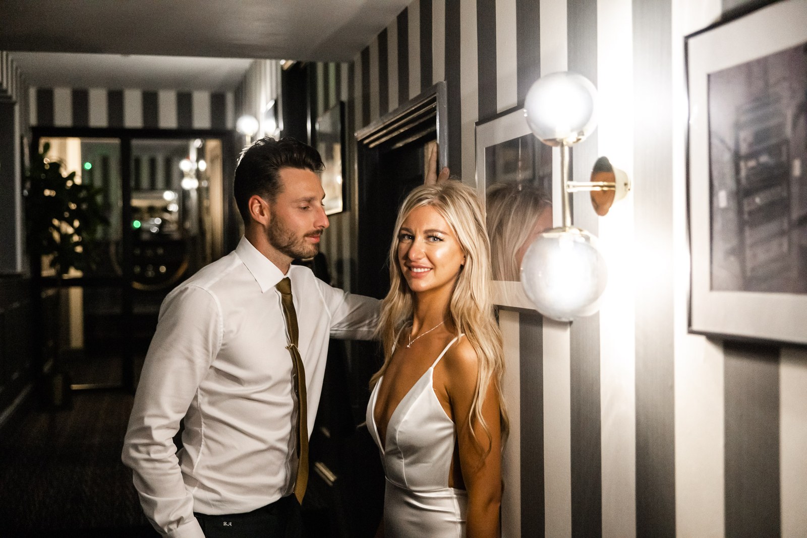 The couple pausing by a wall light in the striped corridor