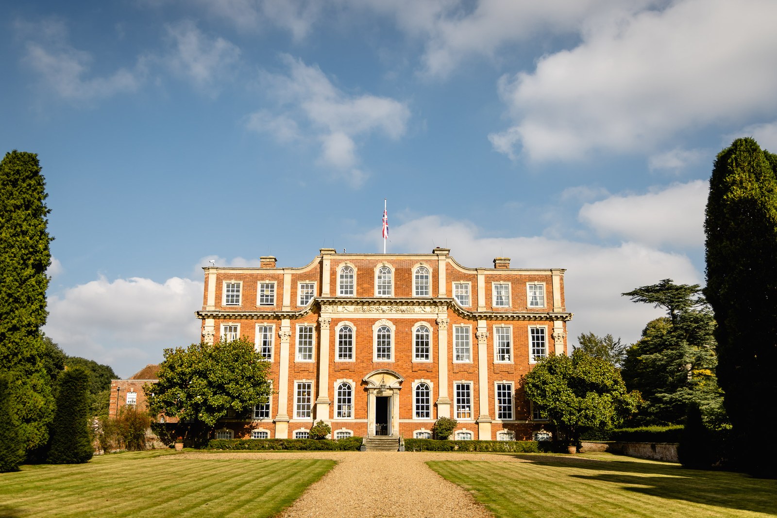 The red-brick Georgian frontage of Chicheley Hall, framed by lawns and a gravel drive under a blue sky