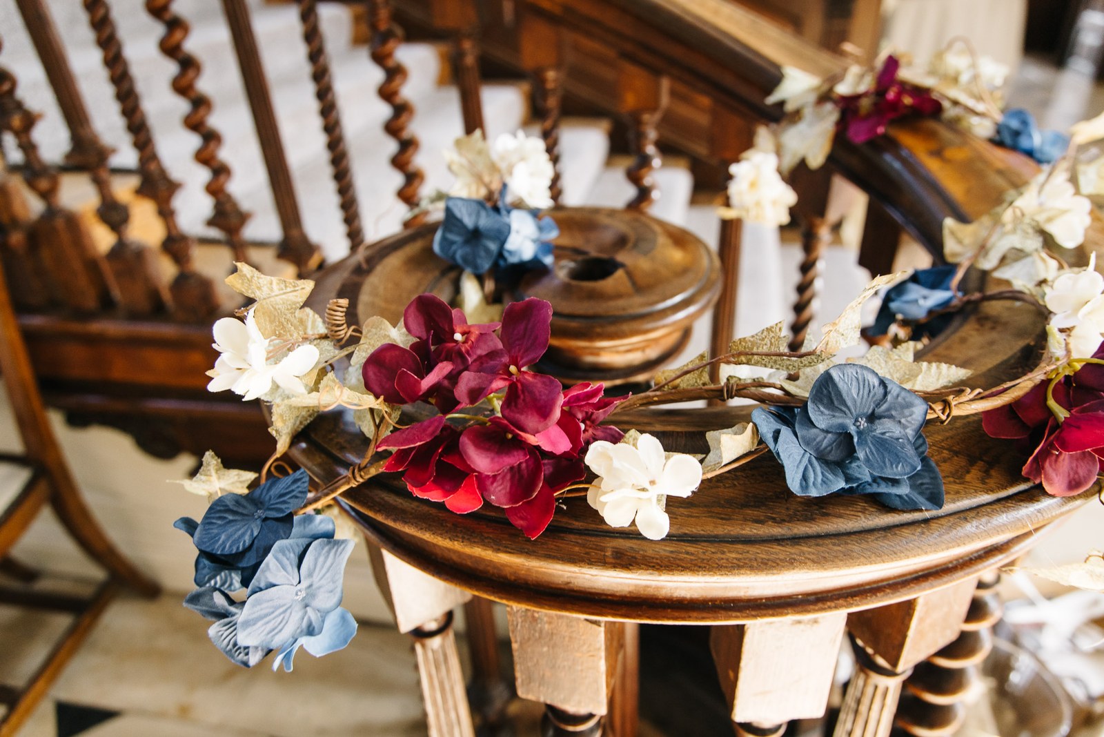 Red and blue floral garland draped over a round wooden table beside a carved staircase