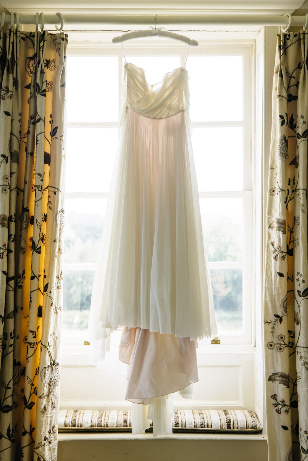 The bride's gown hanging in a tall sash window with patterned curtains