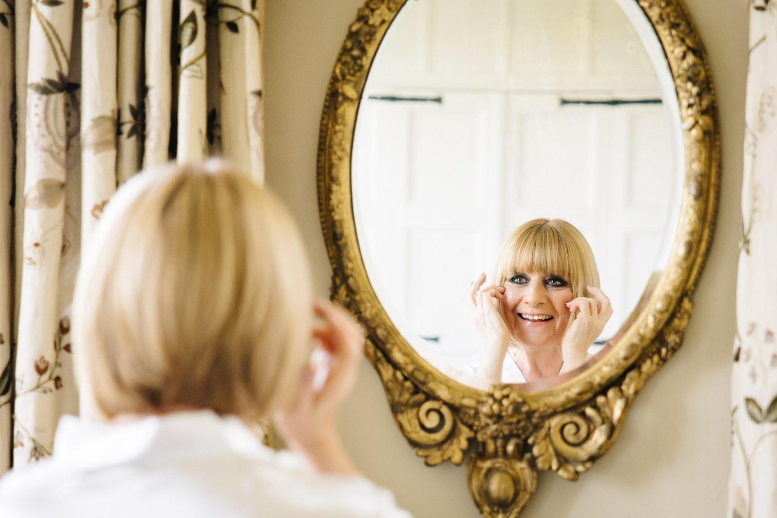 Bride laughing at her reflection in an ornate gilt oval mirror