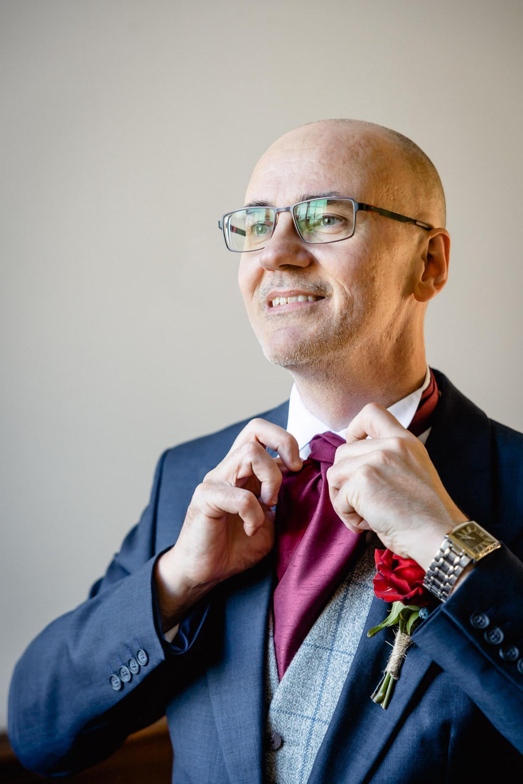 Groom adjusting his burgundy tie, wearing a navy suit with a red rose buttonhole