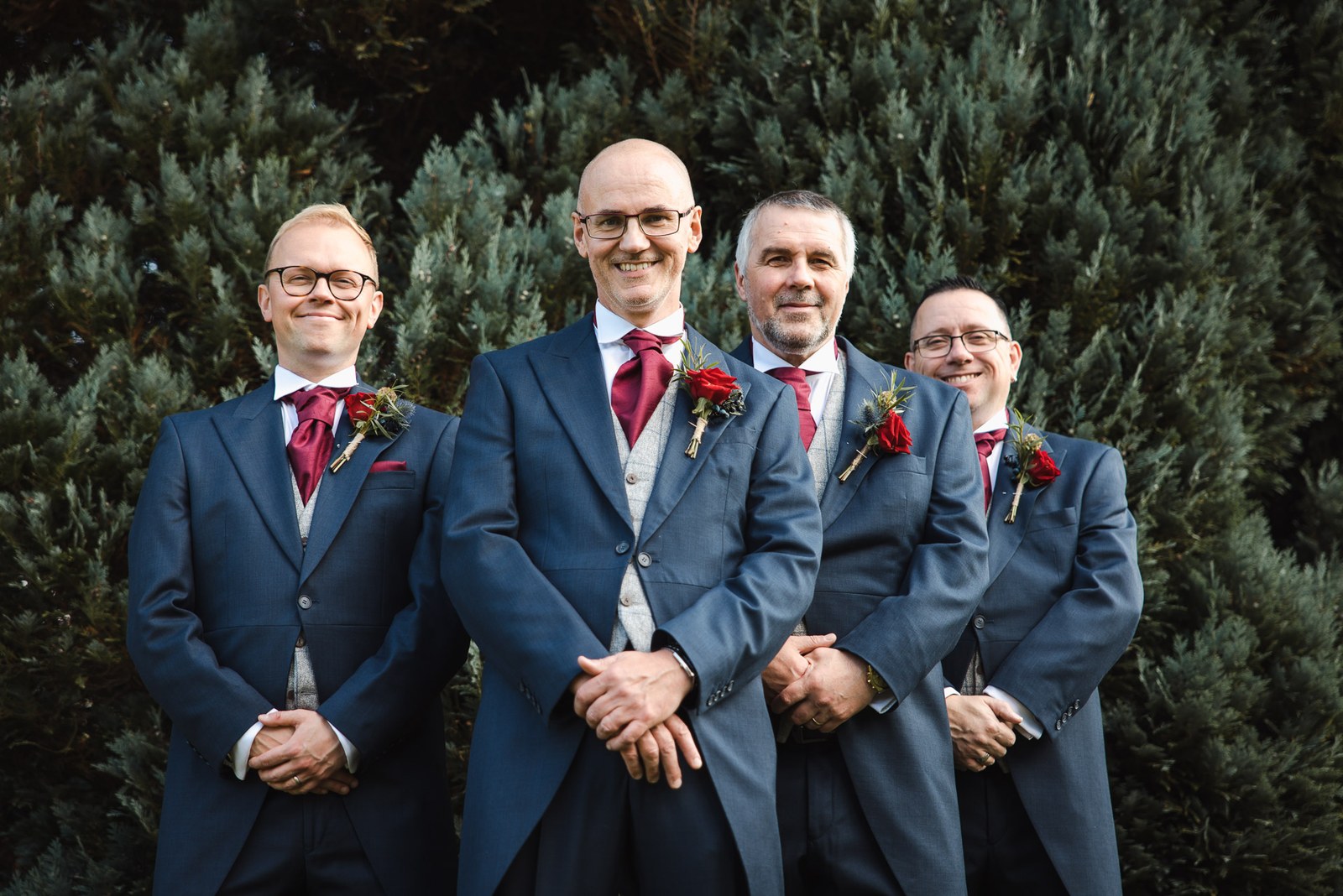 Groom and groomsmen in navy morning suits and burgundy ties beside a clipped hedge