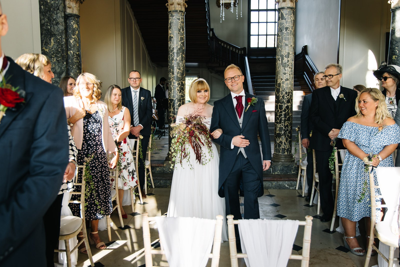 Bride walking down the aisle on her escort's arm past guests in the marble-columned hall