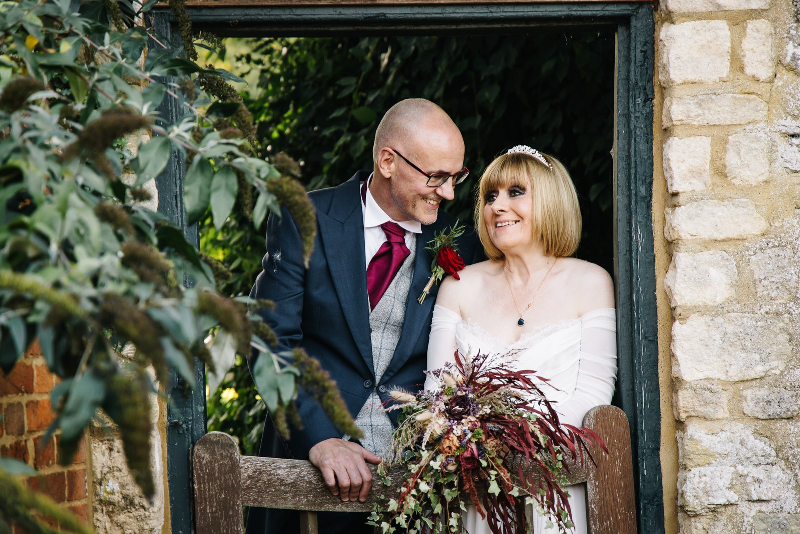 The newlyweds smile together on a bench framed by greenery