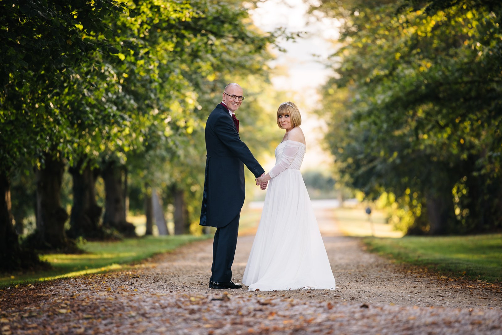 Bride and groom hold hands on a tree-lined avenue scattered with autumn leaves
