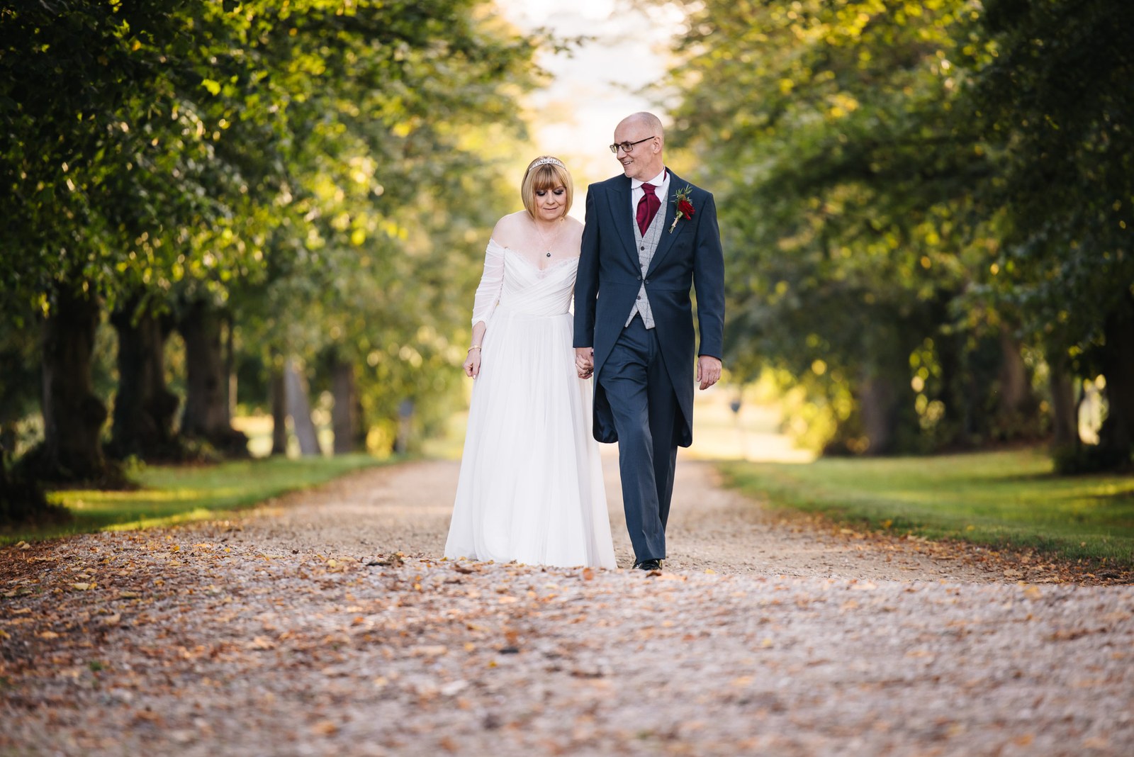 The couple stroll down a tree-lined gravel avenue in golden evening light