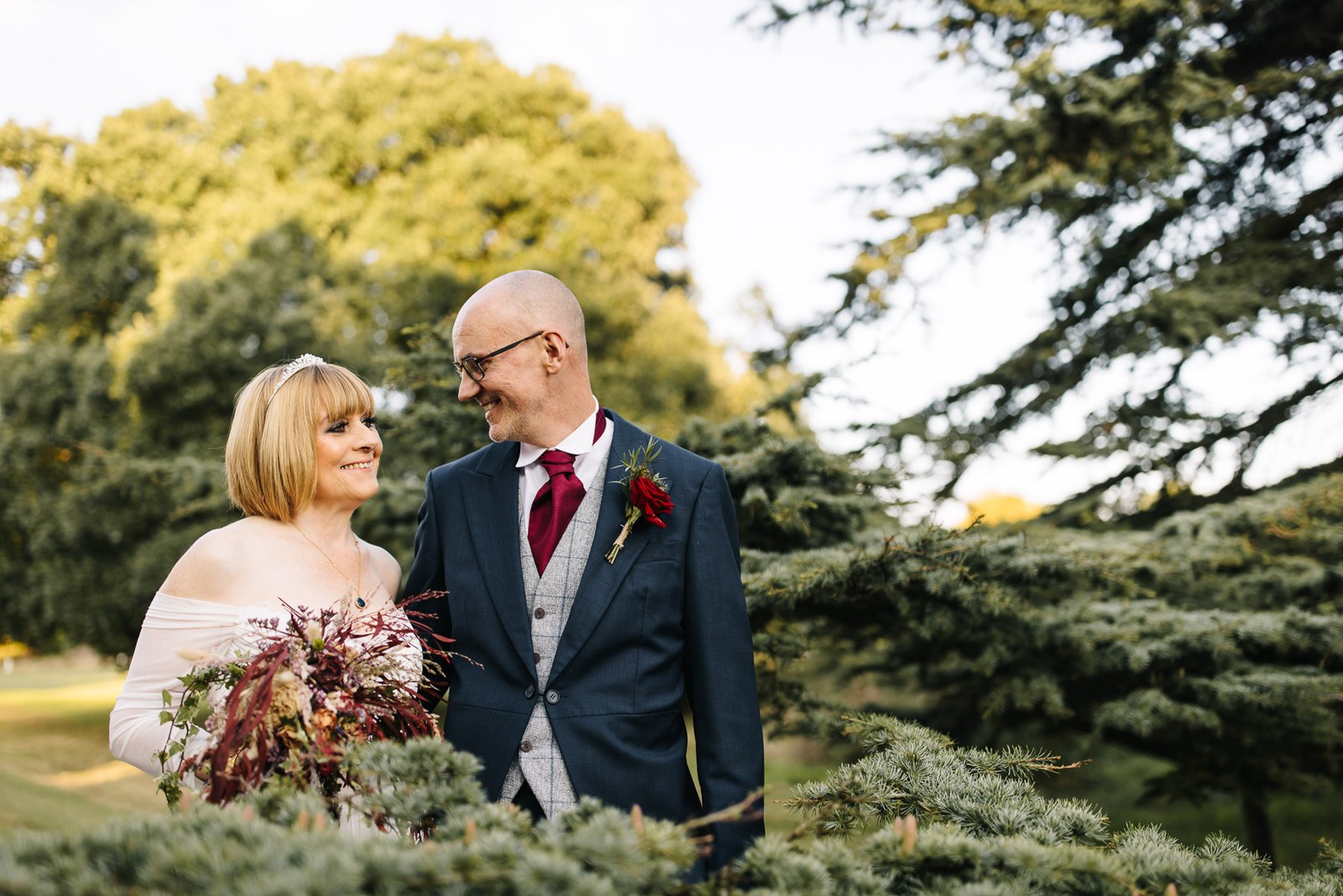 The couple stand among tall cedar trees, the bride holding her autumnal bouquet