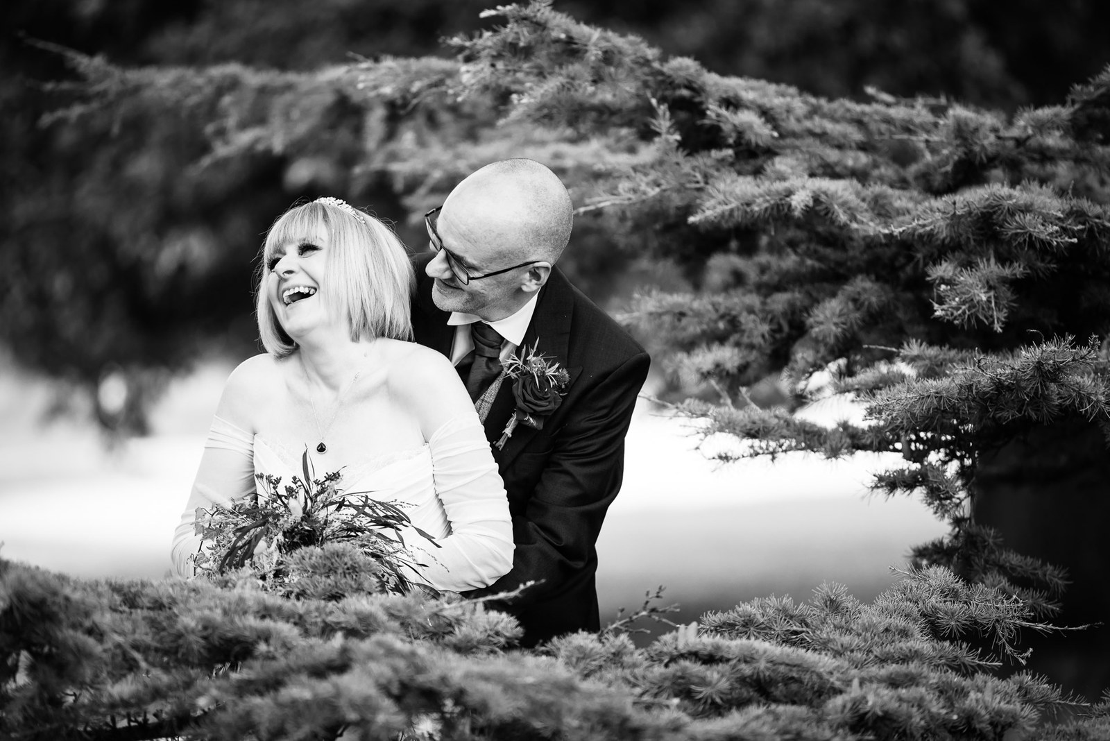 Black-and-white portrait of the couple laughing beneath pine branches