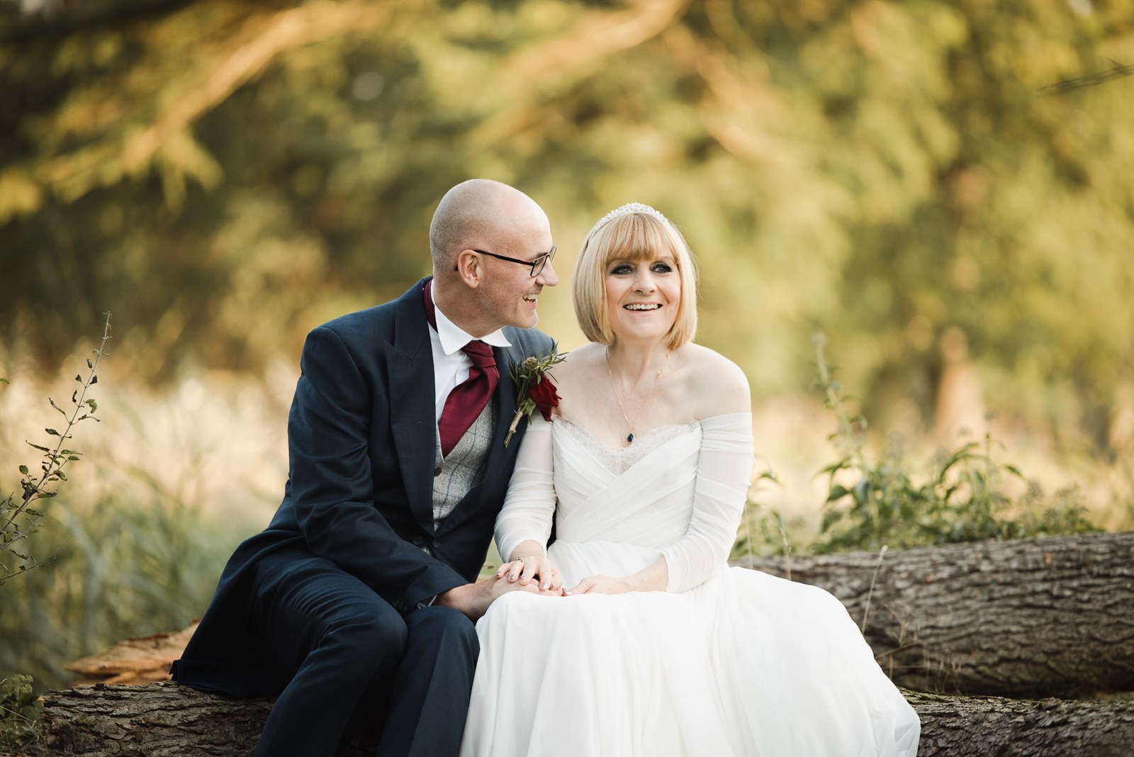 Bride and groom rest on a log beneath trees in warm golden light