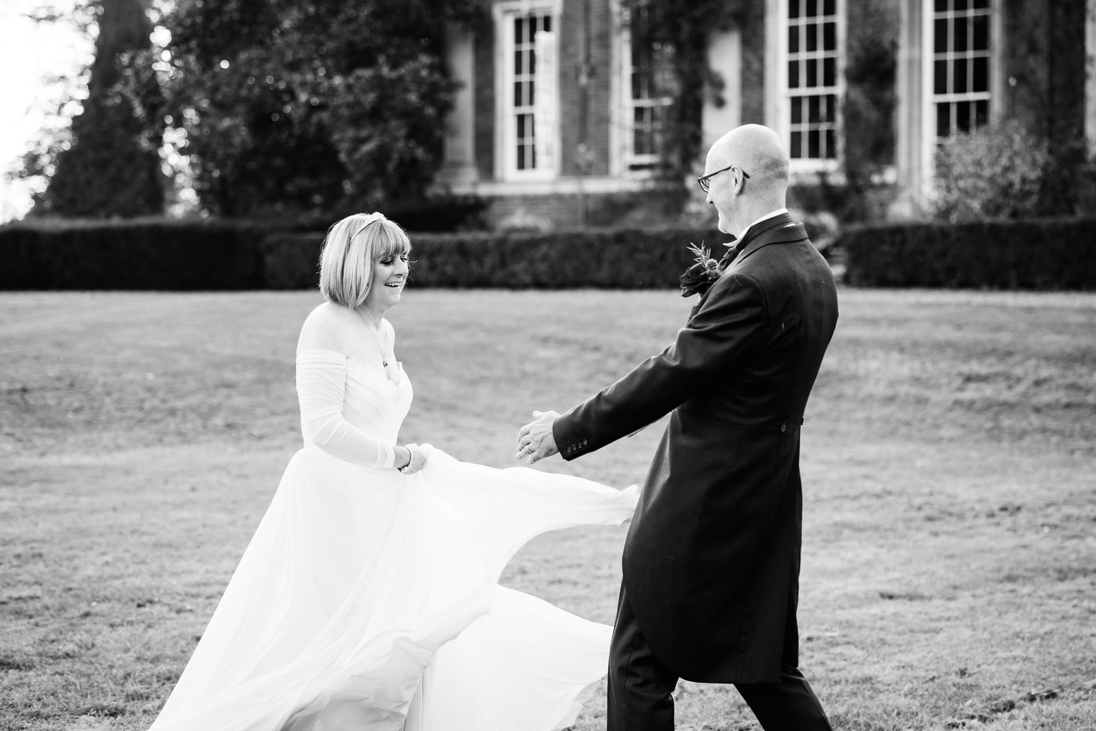 Black-and-white shot of the bride swirling her gown on the lawn as the groom reaches out