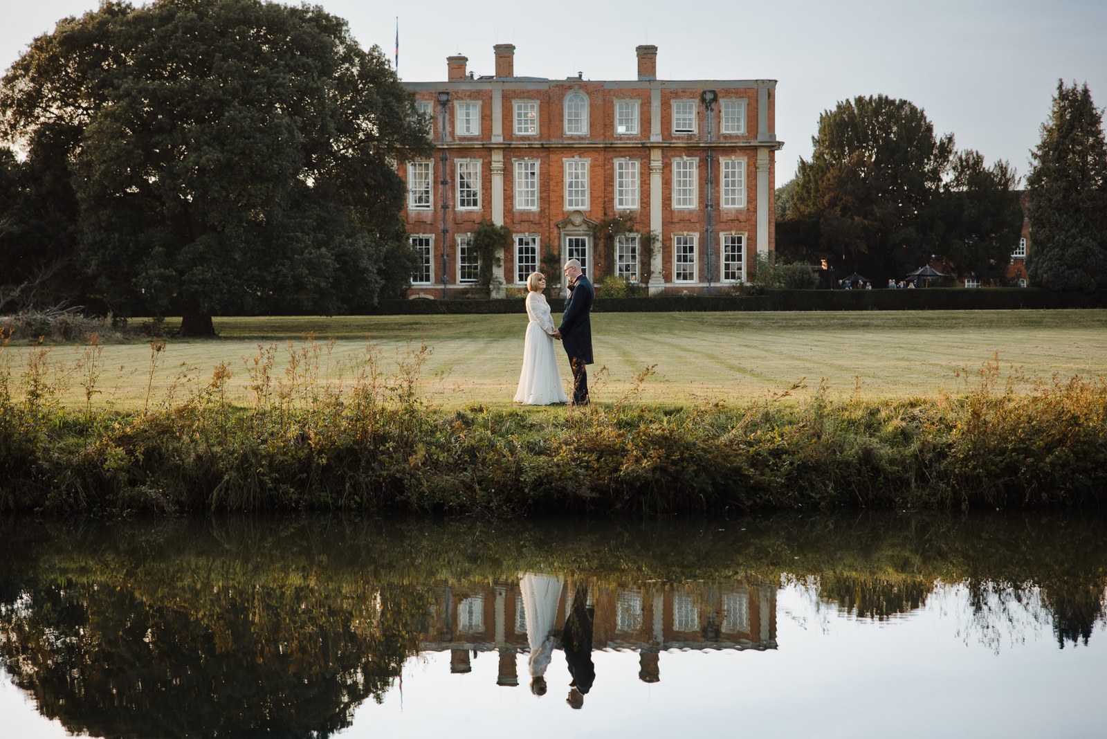 The couple mirrored in the lake before Chicheley Hall at golden hour